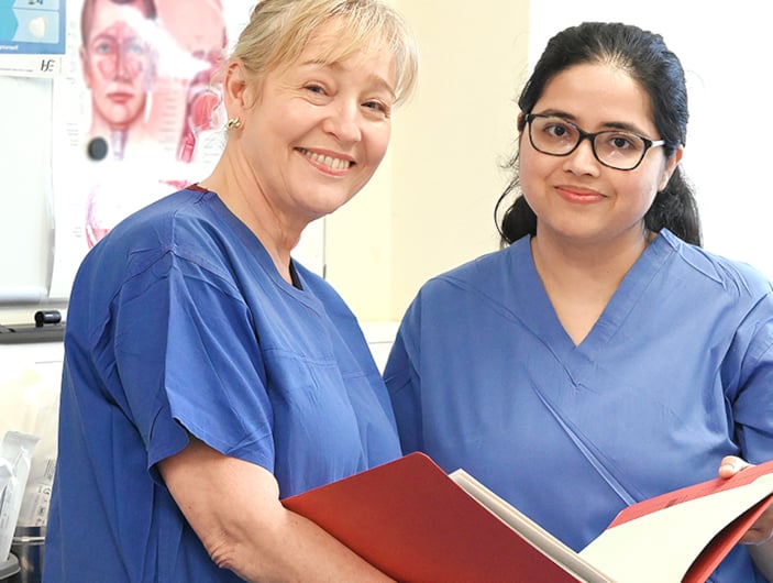 Two nurses in blue scrubs holding an open file look at the camera